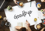 White poster-board sitting on a wooden table features the word 'Goodbye' in black. Coworkers sit on the outskirts of the poster-board with cappuccino, tea, and snacks.