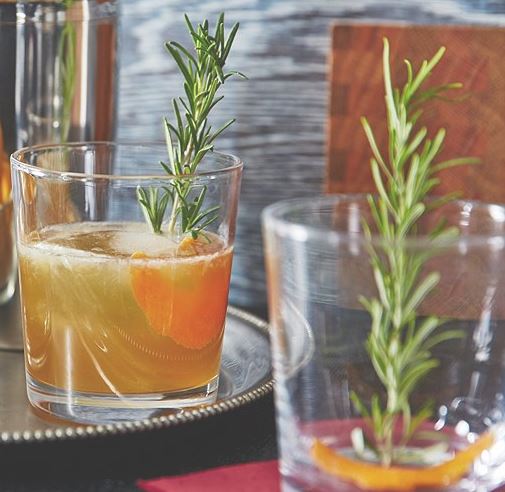 Two glasses with rosemary springs on a bar top, and one is filled with ice, light brown liquid and orange.