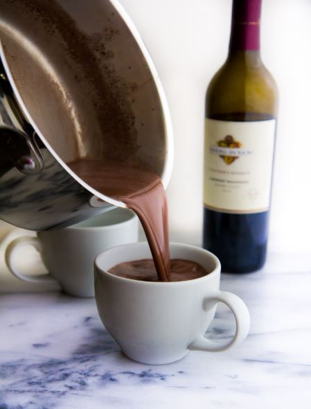 Pouring homemade hot chocolate from a pot into a white mug in front of a bottle of red wine.
