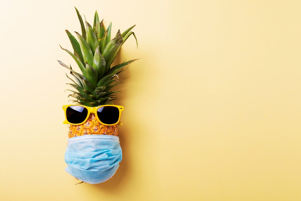 Concept of summer vacation 2020. Pine apple with a medical mask and sun glasses on the yellow background. Top view image