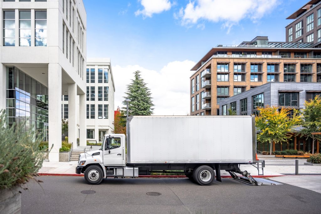 White middle class relocation rig day cab semi truck tractor with long spacious box trailer standing on the urban city street with multilevel apartment and office buildings unloading delivered goods