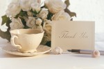 A thank you card, a tea cup and a bouquet on a white table cloth.