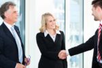 Two men and one woman, outside, dressed in business attire shaking hands.