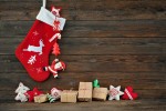 A Christmas stocking hanging with santa hanging from the top and presents below. The background is wooden.