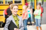 A woman in the foreground in a gray sportcoat holding a black planner with children and a schoolbus in the background.