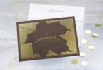 A brown greeting card with a gold foil leaf design on top of a white envelope on a gold-speckled background.